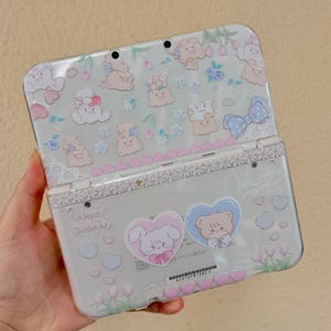 May include: A light gray Nintendo 3DS console with a decorative cover featuring cartoon bears, bunnies, and floral patterns. The cover includes the text "Happy Birthday" and heart-shaped stickers with cute animal designs. The overall design is pastel and whimsical.