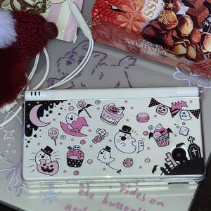 May include: A white Nintendo DS Lite console with a Halloween design featuring ghosts, cupcakes, and a crescent moon. The design includes pink and black accents. The console is on a patterned surface with text that reads "Rides on the butterfly and floats about."