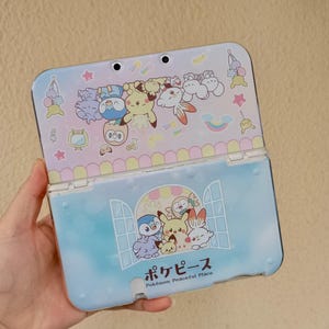 Puede incluir: Una funda azul y blanca para Nintendo 3DS con personajes de dibujos animados. La funda está abierta, revelando un interior azul claro con un diseño de ventana y texto japonés. La parte superior tiene varios personajes y colores pastel.