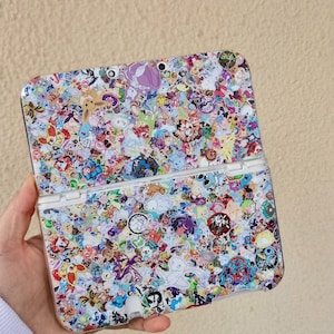 May include: A handheld gaming console with a colorful, patterned design. The console is covered in various cartoon character stickers in a variety of colors. The console is open, revealing the screen and buttons.