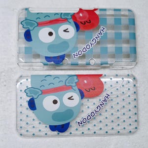 Peut inclure: Deux coques de téléphone transparentes avec un personnage de dessin animé bleu, un bandeau rouge et un œil qui cligne de l'œil. Les coques ont un fond à carreaux et à pois bleus et blancs, avec le texte "HANGYODON" en bleu.