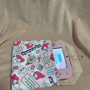 May include: Pink handheld gaming device in a pink and white patterned case featuring My Melody characters. The case has a button closure and the text "My Melody" and "Tea time is my favourite time of the day!"