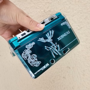 Może przedstawiać: Turkusowa konsola do gier przenośna Nintendo 3DS z przezroczystym etui ochronnym. Konsola zawiera napis "XERNEAS" i białą grafikę jelenia. Na dole konsoli widnieje napis "NINTENDO 3DS".