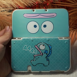 Peut inclure: Une coque de Nintendo 3DS turquoise avec un motif de monstre de dessin animé. La partie supérieure présente de grands yeux et une bouche, tandis que la partie inférieure montre un monstre hurlant avec le mot "AAAA". La coque est ouverte.
