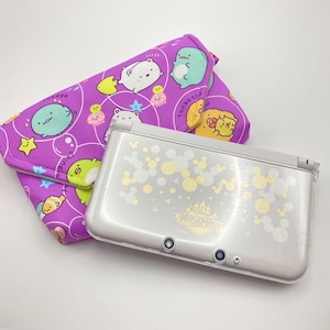 以下が含まれることがあります： 装飾的なデザインのシルバーのニンテンドー3DS携帯ゲーム機と、紫色の布製ケース。ケースには、さまざまな色の漫画のキャラクターの繰り返しパターンがあります。コンソールには、ミッキーマウスの形の装飾的なデザインがあります。