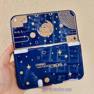 May include: A dark blue protective case for a New Nintendo 3DS XL console. The case features a gold celestial design with stars, planets, and a sleeping character on a crescent moon. The Japanese text "ねむたい" is printed on the top left corner.