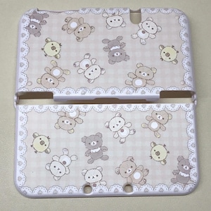 May include: A white and beige Nintendo 3DS case with a repeating pattern of cartoon bears and chicks. The case has a decorative border with a heart and lace design.