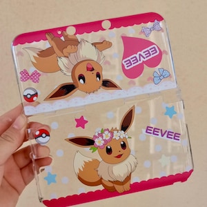 May include: Clear plastic Nintendo DS case featuring Eevee characters. The case has a pink border with white polka dots and decorative elements like bows, stars, and a heart with the word "EEVEE". The case is designed to protect the Nintendo DS.