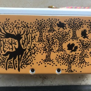 May include: A tan handheld gaming device with a black forest design featuring a deer and birds. The cover has a white top and two circular holes. The design is printed on the device's cover.