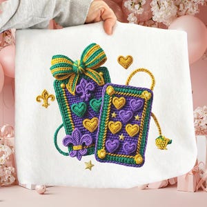 May include: A white fabric with a colorful embroidered design featuring a bow, fleur-de-lis, and two rectangular shapes. The shapes are decorated with hearts and stars in shades of purple, green, and gold.