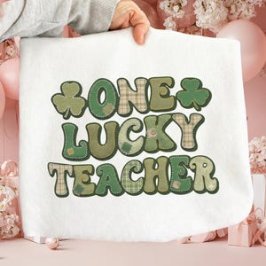 May include: White fabric with the text "ONE LUCKY TEACHER" in green and tan patchwork letters. The design includes shamrocks. The fabric is held up by a person's hand.