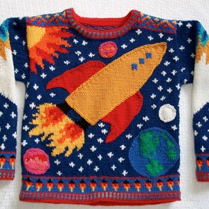 Könnte beinhalten: Ein blauer Strickpullover mit einer Rakete, Planeten und Sternen. Die Rakete ist rot, orange und gelb. Die Planeten sind grün, blau und rosa. Die Sterne sind weiß.