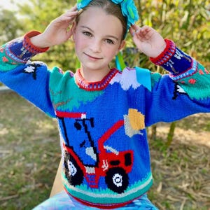 May include: A long-sleeved knit jumper featuring a vibrant blue base with a prominent red tractor graphic on the front. The design includes green landscape accents, a yellow sun, and a red and white striped collar, cuffs, and hemline detail.