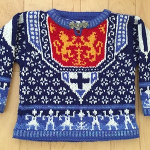 May include: A blue knitted jumper with a white and blue snowflake pattern. The jumper has a red, yellow, and orange crest with two lions on the front. The jumper has a silver clasp at the neckline.
