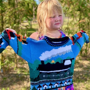 May include: A long-sleeve knit sweater featuring a vibrant blue background with a detailed graphic of a black steam locomotive train emitting white smoke. The design includes green hills, a small yellow tree, and a row of white houses along the hem.