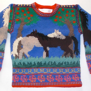 Peut inclure: Un pull en tricot bleu avec un motif coloré de chevaux et d'arbres. Le pull a une bordure rouge, blanche et bleue autour du cou et des poignets.