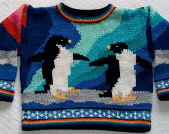 May include: A blue knitted jumper with a rainbow stripe around the neck and a penguin design. The penguins are black and white with orange beaks and feet. The jumper has a white and blue snowflake pattern on the cuffs and hem.