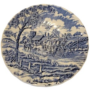 May include: A round, off-white plate with a blue transferware design. The scene depicts a horse-drawn carriage on a road, with a church and trees in the background. The plate has a scalloped edge.