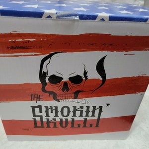 May include: A white box with red and blue stripes and stars. The box has a skull with a cigar in its mouth and the text "The Smokin' Skull" on it.