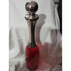 May include: A tall, red glass bottle with a silver top and a round, silver knob. The bottle has a unique, curved shape.