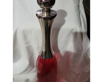 May include: A tall, red glass bottle with a silver top and a round, silver knob. The bottle has a unique, curved shape.