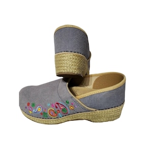 May include: A pair of grey canvas clogs featuring a woven jute-style platform sole and beige trim. The exterior of the shoes is decorated with colorful embroidered floral and paisley patterns in shades of pink, blue, green, and orange.