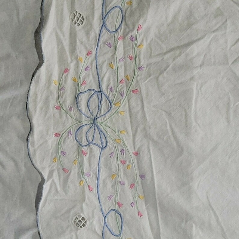 K&ouml;nnte beinhalten: Wei&szlig;er Stoff mit einem gewellten Rand und einem gestickten blauen Band mit rosa, gelben und gr&uuml;nen Blumen.