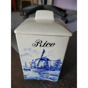 May include: A white ceramic rice container with a lid and a blue and white windmill design. The word "Rice" is written in black script on the front of the container. The container has a rectangular shape.