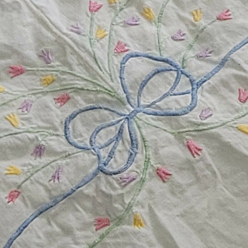 K&ouml;nnte beinhalten: Eine wei&szlig;e Stofftischdecke mit einer blauen, gestickten Schleife in der Mitte. Die Schleife ist von gestickten Blumen in Rosa-, Gelb- und Gr&uuml;nt&ouml;nen umgeben.