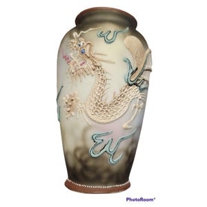 May include: A frosted glass vase featuring a raised, textured dragon motif in cream and white. The design includes accents of light blue and pink, with a dark, smoky gradient at the base and a solid brown rim at the top and bottom edges.