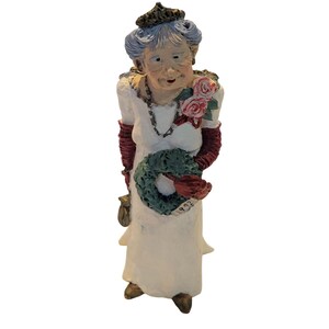 May include: A white porcelain figurine of a woman wearing a white dress, a red and gold crown, and red gloves. She is holding a green wreath and a bouquet of pink roses. The figurine has the number 1992 on the base.