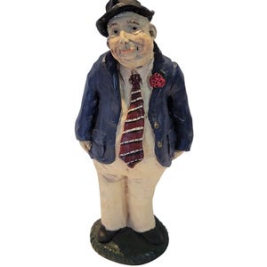 May include: A ceramic figurine of a man in a blue blazer, white shirt, and striped tie. He wears a black hat and has a red flower on his lapel. The figure stands on a green base.