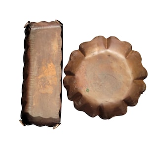 May include: Two antique copper items: a rectangular box with a scalloped edge and a round, flower-shaped dish. The box has a detailed design on its surface. Both items have a warm, aged patina, suggesting they are vintage or vintage-inspired.