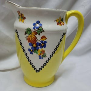May include: A vintage ceramic pitcher with a yellow base and handle, and a white upper section. The white section features a floral design with blue, yellow, and orange flowers. A black and white checkered pattern borders the floral design.