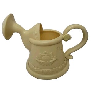 May include: Cream-colored watering can with a curved spout and handle. The body features a floral design and decorative trim. The watering can is made of a solid material and is designed for gardening.
