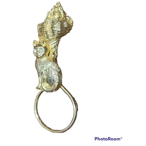 May include: A gold-toned cat-shaped brooch with a textured finish. The cat has rhinestone eyes and sits below a textured, fan-shaped element. A circular ring is attached below the cat. The brooch is likely vintage.