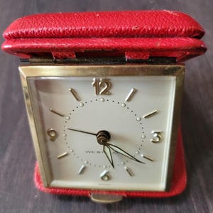 May include: A vintage travel alarm clock with a red leather case. The clock face is white with gold-colored hands and hour markers. The clock is marked "WESTCLOX" and "GERMANY". The case is folded open, revealing the clock face.