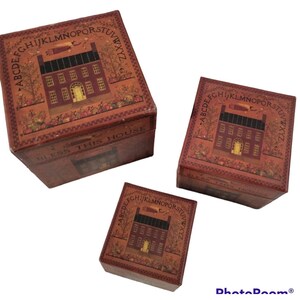 May include: Three square boxes in varying sizes, each featuring a dark red finish with a printed illustration of a house, an alphabet border, and the text BLESS THIS HOUSE. The design includes trees, foliage, and a flying figure.