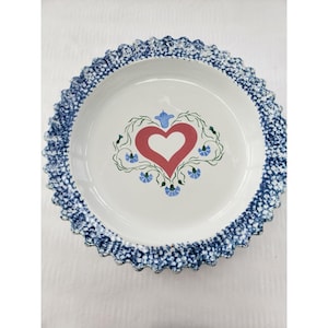May include: White ceramic pie plate with a blue dotted scalloped edge. The center features a red heart design with green vines and blue floral accents. A charming addition to any kitchen.