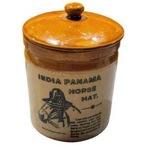 May include: A vintage, cylindrical stoneware jar with a lid. The jar is tan with brown accents and features the text "INDIA PANAMA HORSE HAT" along with an illustration of a horse wearing a hat. The lid has a small knob.