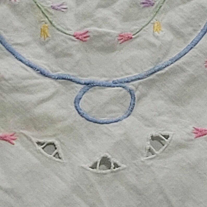 K&ouml;nnte beinhalten: Wei&szlig;e Baumwolltischdecke mit blauen und rosa gestickten Blumen und einem blauen Kreis in der Mitte. Die Tischdecke hat ausgeschnittene Designs an den R&auml;ndern.