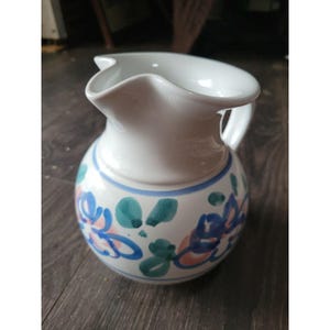May include: White ceramic pitcher with a rounded body and a wavy rim. The pitcher is decorated with blue, pink, and green floral patterns and a blue stripe near the base. It has a handle on the side.