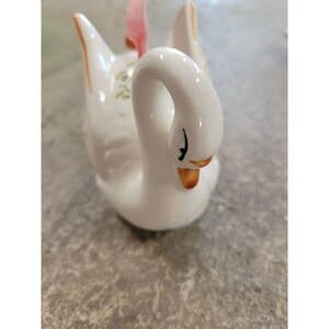 May include: A white ceramic swan figurine with a curved neck and orange beak. The swan has black eyes and pink accents on the wings. Delicate floral details are visible on the back. A decorative piece.