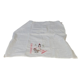 May include: A white cloth with a Southwestern-themed embroidered design. The embroidery features a candle, a cactus, and other shapes in red, black, and brown. The cloth is folded and appears to be a kitchen towel or napkin.