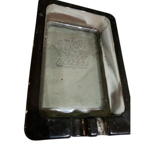 May include: Rectangular ashtray with a black frame and a silver interior. The interior features an embossed design with the word "Garage" and a stylized emblem. The ashtray has a small notch for resting cigarettes.