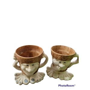 May include: Two ceramic mugs shaped like faces with brown and white glaze. The mugs have a whimsical, cartoon-like appearance with exaggerated features. The mugs are sitting on a white surface.