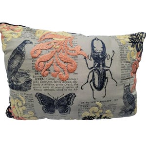 May include: A rectangular throw pillow with a light gray background featuring printed black text from an encyclopedia, black illustrations of a bird, a beetle, a butterfly, and an owl, and embroidered floral patterns in peach and cream thread.