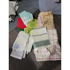 May include: A collection of folded fabric linens including white cloths with floral embroidery, a light green cloth with leaf patterns, a tan cloth with a basket design, a white cloth with a green geometric border, and a white cloth with red grid lines.