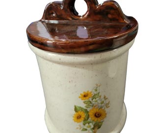 Könnte beinhalten: Ein wandmontierter Salztopf aus Keramik mit einem gesprenkelten cremeweißen Körper und einem dunkelbraun glasierten Deckel. Die Vorderseite zeigt ein Blumenmotiv mit gelben Sonnenblumen und kleinen weißen Blüten. Der obere Teil hat einen Griff.