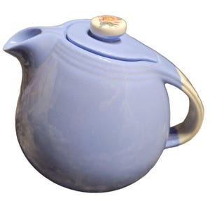 May include: A light blue ceramic teapot with a rounded body and a curved spout. The handle is off-white, and the lid features a floral design on the knob. The teapot is designed for serving tea.